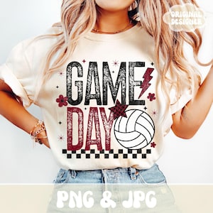 Puede incluir: Camiseta color crema con las palabras "GAME DAY" en negro y burdeos, con un gráfico de voleibol. El diseño incluye un rayo, estrellas y detalles florales. La camiseta es una prenda informal.