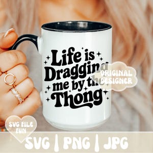 May include: White ceramic mug with a black interior and handle. The mug features the text "Life is Dragging me by the Thong" in black, with star accents. A fun and quirky design for coffee or tea.