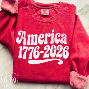 May include: A red sweatshirt with the word "America" in large white letters, along with the numbers "1776-2026". The sweatshirt has a crew neck and long sleeves with a soft, pinkish lining. The text "DO NOT COPY" is also visible.