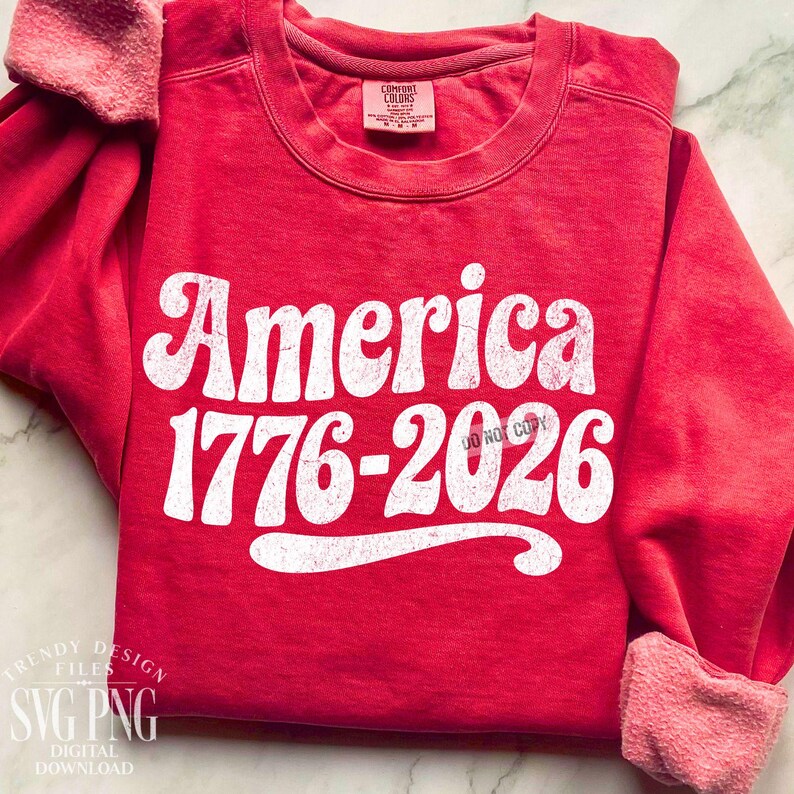 May include: A red sweatshirt with the word "America" in large white letters, along with the numbers "1776-2026". The sweatshirt has a crew neck and long sleeves with a soft, pinkish lining. The text "DO NOT COPY" is also visible.