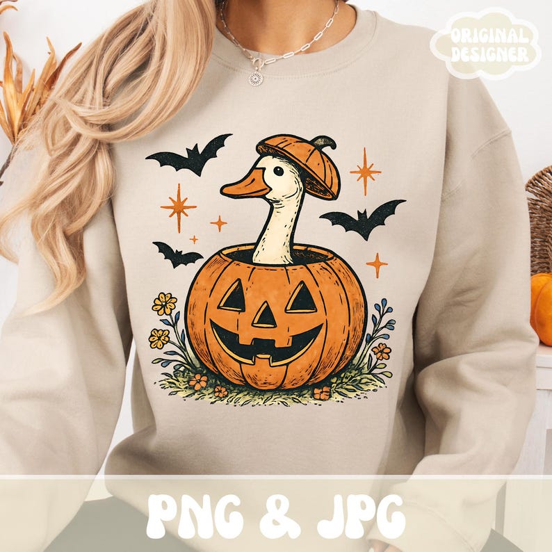 May include: Beige sweatshirt with a Halloween-themed graphic. The design features a white goose wearing a pumpkin hat, emerging from a carved orange pumpkin. Bats and stars surround the goose and pumpkin. The text "PNG & JPG" is at the bottom.