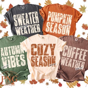 Op de afbeelding: Vijf T-shirts in verschillende kleuren, elk met een herfstthema zin in versleten witte letters. Zinnen zijn onder meer "Sweater Weather", "Pumpkin Season", "Autumn Vibes", "Cozy Season" en "Coffee Weather". De shirts zijn omgeven door herfstbladeren.