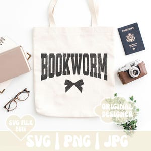 May include: A white tote bag with black text that reads "BOOKWORM" and a black bow. The bag is on a white surface with a passport, a camera, and a plant.