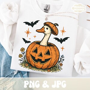 May include: White sweatshirt featuring a Halloween-themed graphic. The design includes a goose wearing a pumpkin hat, emerging from a carved pumpkin. Bats, stars, and flowers surround the pumpkin. The text "PNG & JPG" is at the bottom.