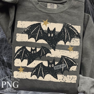 May include: A charcoal gray crewneck sweatshirt featuring a graphic print of three black bats flying across horizontal, distressed beige stripes. The design includes small gold stars scattered between the bats. The collar tag reads Comfort Colors.