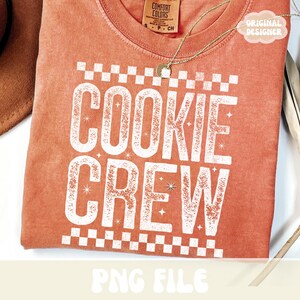 May include: A coral-colored t-shirt with the words "COOKIE CREW" in large, white, distressed lettering. The design includes a checkered border and star accents. A gold necklace and a tag that says "ORIGINAL DESIGNER" are visible.