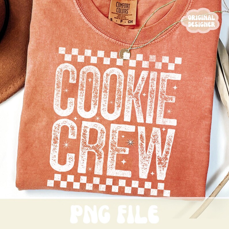 May include: A coral-colored t-shirt with the words "COOKIE CREW" in large, white, distressed lettering. The design includes a checkered border and star accents. A gold necklace and a tag that says "ORIGINAL DESIGNER" are visible.