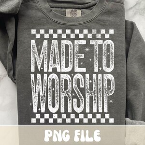 K&ouml;nnte beinhalten: Ein dunkelgraues Sweatshirt mit dem Schriftzug "MADE TO WORSHIP" in gro&szlig;en, verwaschenen wei&szlig;en Buchstaben. Der Text wird von einem Schachbrettmuster umrahmt. Die Marke "Comfort Colors" ist oben sichtbar. Der untere Teil des Bildes zeigt "PNG FILE".