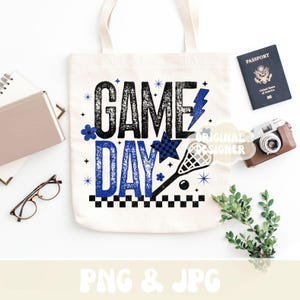 May include: A white tote bag with the words "GAME DAY" in black and blue, along with a lacrosse stick graphic. The bag is surrounded by a passport, camera, glasses, and a notebook.