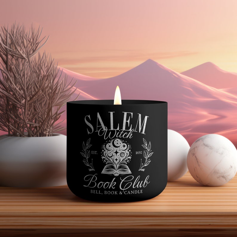 K&ouml;nnte beinhalten: Schwarze Kerze mit einem wei&szlig;en Design, auf dem "Salem Witch Book Club" steht, mit einer hexenhaften Illustration eines Buches und Sternen. Der Text lautet au&szlig;erdem "Est. 1692" und "Bell, Book & Candle".