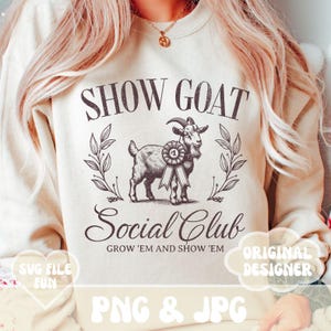 May include: A beige sweatshirt with a black and white illustration of a goat wearing a ribbon. The text "SHOW GOAT Social Club GROW 'EM AND SHOW 'EM" is printed on the sweatshirt. The text "ORIGINAL DESIGNER" is in a speech bubble on the right side of the sweatshirt. The text "SVG FILE FUN PNG & JPG" is printed at the bottom of the sweatshirt.