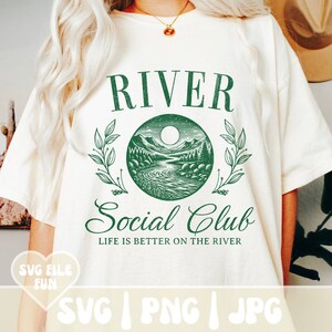 May include: Cream-colored t-shirt with a green graphic. The graphic features the words "RIVER Social Club" above and below a circular illustration of a river scene with mountains, trees, and a moon. The text "LIFE IS BETTER ON THE RIVER" is below the graphic.
