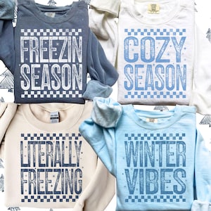 May include: Four crewneck sweatshirts in various colors: dark gray, white, beige, and light blue. Each sweatshirt features a winter-themed phrase in a distressed font: "FREEZIN SEASON", "COZY SEASON", "LITERALLY FREEZING", and "WINTER VIBES".