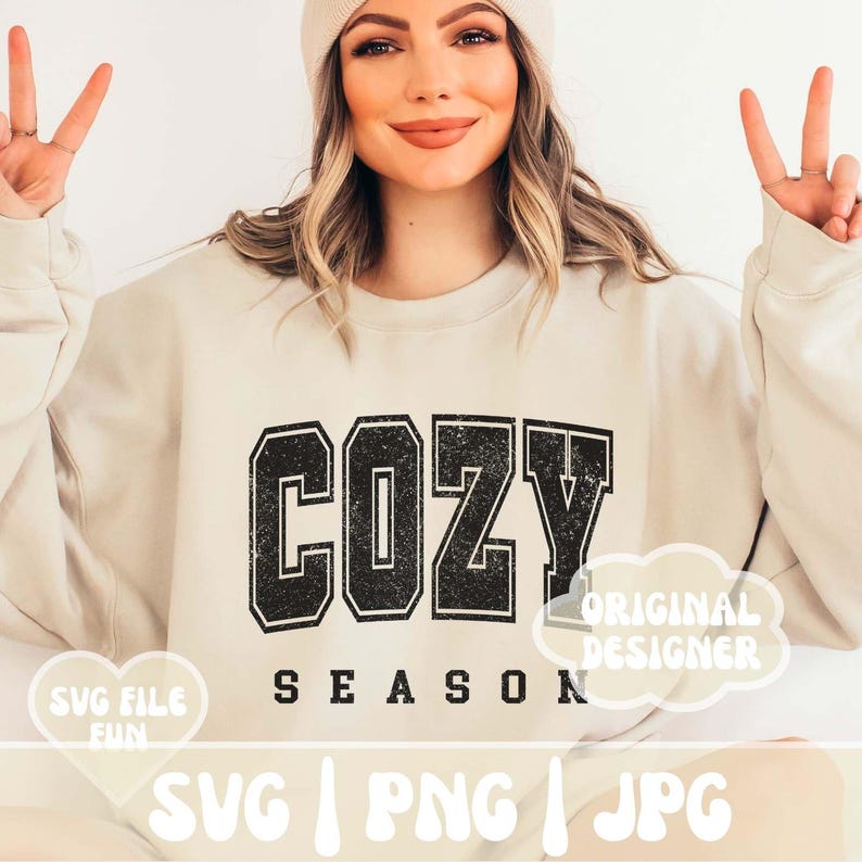 May include: A beige sweatshirt with the text "Cozy Season" in black with a distressed effect. The text is in a large, bold font. The sweatshirt is being worn by a person with a white beanie. The image also includes the text "Original Designer" in a white cloud shape.