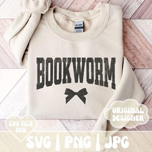 May include: A beige sweatshirt with the text "BOOKWORM" in black with a distressed look. A black bow is below the text. The sweatshirt is on a wooden surface.