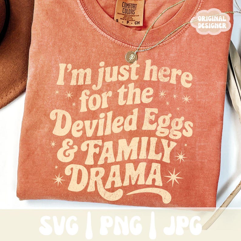 Puede incluir: Una camiseta de color coral con el texto "I'm just here for the Deviled Eggs & FAMILY DRAMA" en una fuente retro. La camiseta tiene cuello redondo y mangas cortas. Se ve un collar dorado con un colgante.
