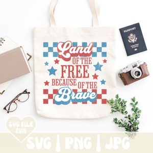 May include: Cream-colored tote bag with the text "Land of the Free Because of the Brave" in red and blue. The design includes stars and a checkered pattern. A passport, camera, and other items are arranged around the bag.