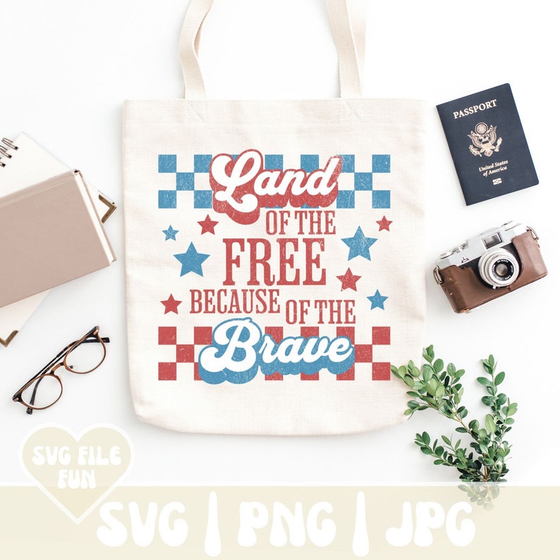 May include: Cream-colored tote bag with the text "Land of the Free Because of the Brave" in red and blue. The design includes stars and a checkered pattern. A passport, camera, and other items are arranged around the bag.