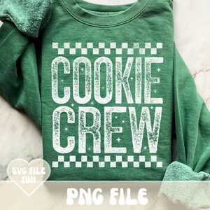 May include: Green sweatshirt with the words "COOKIE CREW" in large, white, distressed lettering. The design includes a checkered pattern above and below the text, with star accents. The phrase "SVG FILE FUN" and "PNG FILE" are also visible.