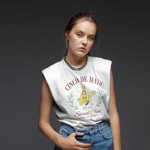 Puede incluir: Camiseta sin mangas blanca con un dise&ntilde;o de Cinco de Mayo. El dise&ntilde;o incluye una botella de tequila, una margarita y el texto "Social Club" y "Nacho Average Fiesta". La modelo lleva un collar de cadena plateada y vaqueros azules.