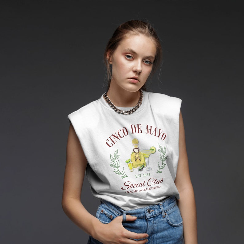 Puede incluir: Camiseta sin mangas blanca con un dise&ntilde;o de Cinco de Mayo. El dise&ntilde;o incluye una botella de tequila, una margarita y el texto "Social Club" y "Nacho Average Fiesta". La modelo lleva un collar de cadena plateada y vaqueros azules.
