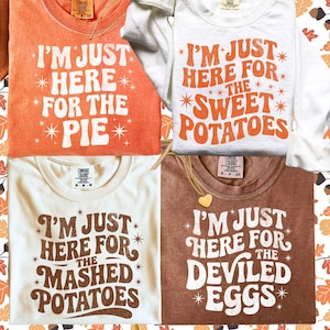 Op de afbeelding: Vier T-shirts in oranje, wit, crème en bruin, elk met een andere voedselgerelateerde zin in witte letters. De zinnen zijn: "I'm Just Here For The Pie", "I'm Just Here For The Sweet Potatoes", "I'm Just Here For The Mashed Potatoes" en "I'm Just Here For The Deviled Eggs."