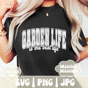 May include: A black t-shirt with white text that reads "GARDEN LIFE is the best life". The text has a distressed, vintage look. The shirt is being worn by a person with long blonde hair.