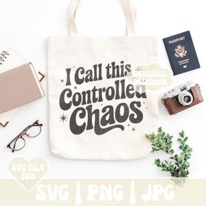 May include: A cream-colored tote bag with the text "I call this Controlled Chaos" in a retro font. The image includes a passport, a vintage camera, glasses, a notebook, and greenery, suggesting a travel or lifestyle theme.