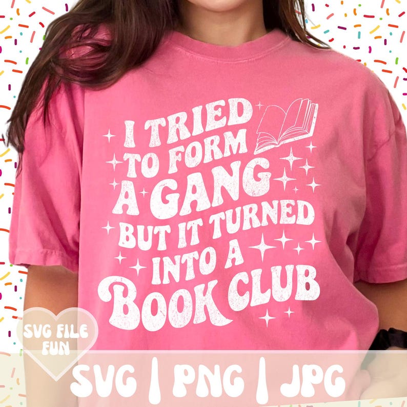May include: Pink t-shirt with the text "I tried to form a gang but it turned into a book club" in white. The design includes an open book graphic and star accents. The shirt is a casual style.
