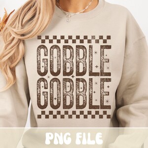 Może przedstawiać: Beżowa bluza z napisem "GOBBLE GOBBLE" w brązowej, postarzanej czcionce. Tekst jest obramowany wzorem w szachownicę. Na dole obrazu widnieje napis "PNG FILE".