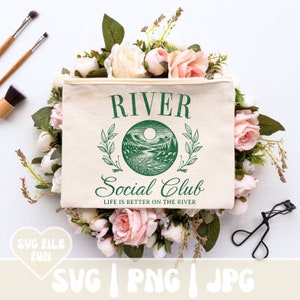 May include: A beige canvas makeup bag with the words "RIVER Social Club" in green. The bag features a circular graphic of a river scene and the text "LIFE IS BETTER ON THE RIVER". The bag is surrounded by pink and white roses and greenery.