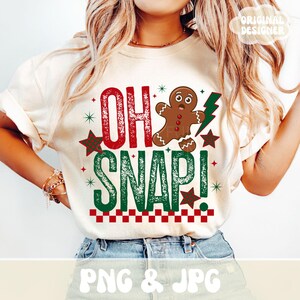 May include: Cream-colored t-shirt with the text "OH SNAP!" in red and green, featuring a gingerbread man and a green lightning bolt. The design includes red and green stars and a checkered border.