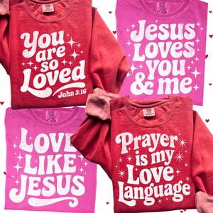 Op de afbeelding: Vier shirts met lange mouwen in rood- en roze tinten. Elk shirt toont witte tekst met zinnen als "You are so Loved" en "Jesus Loves You & Me". De shirts hebben een vintage, retro stijl.