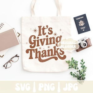 May include: A cream-colored tote bag with the phrase "It's Giving Thanks" in brown lettering. The bag is surrounded by a passport, a camera, a notebook, glasses, and greenery, all on a white surface. The design has a retro aesthetic.