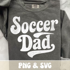 Op de afbeelding: Een donkergrijze sweatshirt met de tekst "Soccer Dad" in grote, witte letters met een distressed look. De sweatshirt heeft een ronde hals en lange mouwen. De tekst "PNG & SVG" staat onderaan de afbeelding.