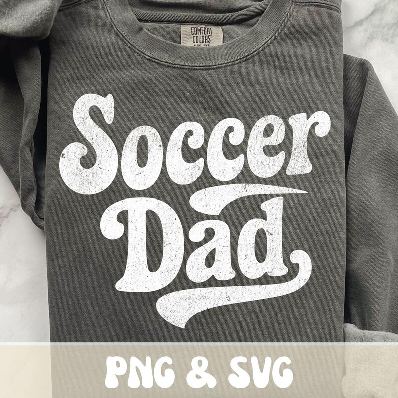 Op de afbeelding: Een donkergrijze sweatshirt met de tekst "Soccer Dad" in grote, witte letters met een distressed look. De sweatshirt heeft een ronde hals en lange mouwen. De tekst "PNG & SVG" staat onderaan de afbeelding.