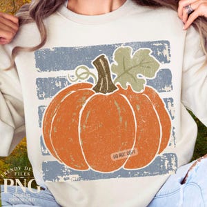 May include: A cream-colored crewneck sweatshirt featuring a large, distressed-style graphic of an orange pumpkin with a brown stem and a light green leaf. The pumpkin is centered over a background of horizontal, weathered blue stripes.