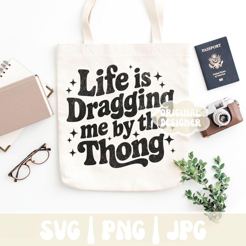 May include: Cream-colored tote bag with the text "Life is Dragging me by the Thong" in black. The bag is surrounded by a passport, a camera, glasses, a notebook, and greenery. The bag is a neutral color.