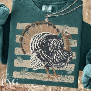 May include: A dark green crewneck sweatshirt featuring a distressed graphic of a turkey with brown and black feathers. The turkey is positioned over horizontal beige stripes and small leaf illustrations. A gold chain necklace rests on the collar.
