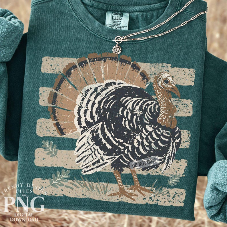May include: A dark green crewneck sweatshirt featuring a distressed graphic of a turkey with brown and black feathers. The turkey is positioned over horizontal beige stripes and small leaf illustrations. A gold chain necklace rests on the collar.