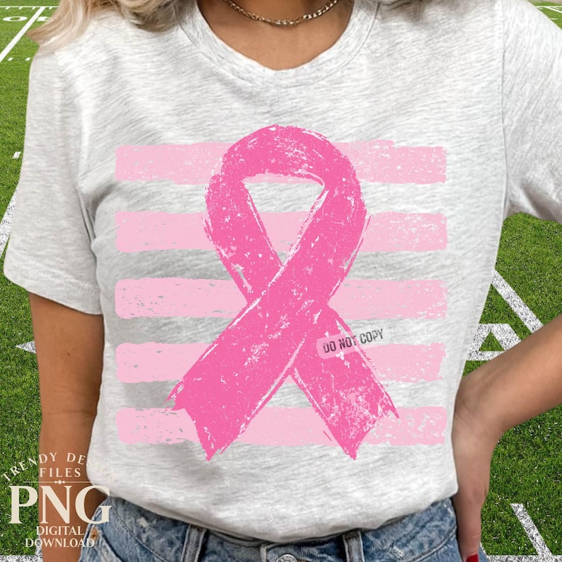 May include: A light gray heathered t-shirt featuring a large, distressed pink awareness ribbon centered over a background of horizontal pink brushstroke stripes. The text DO NOT COPY is printed in small black letters on the ribbon graphic.