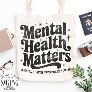 May include: Cream-colored tote bag with the text "Mental Health Matters" in black, along with the phrase "Mental Health Awareness Month." The bag is surrounded by a passport, a vintage camera, and other accessories.