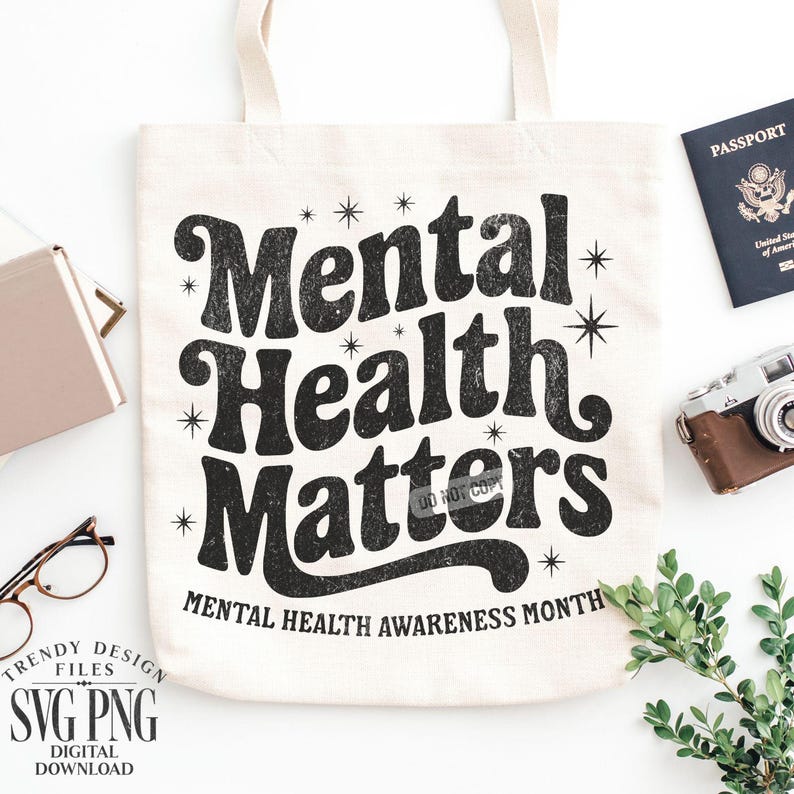 May include: Cream-colored tote bag with the text "Mental Health Matters" in black, along with the phrase "Mental Health Awareness Month." The bag is surrounded by a passport, a vintage camera, and other accessories.