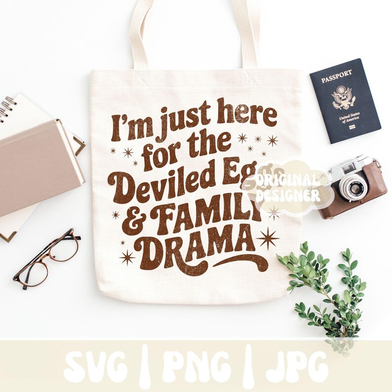 Puede incluir: Bolso tote crema con texto marr&oacute;n: "I'm just here for the Deviled Eggs & FAMILY DRAMA". La imagen incluye un pasaporte, una c&aacute;mara, gafas y vegetaci&oacute;n sobre una superficie blanca.