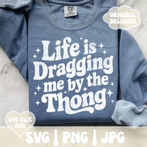 May include: A blue sweatshirt with the phrase "Life is Dragging me by the Thong" in white, retro-style lettering. The design includes star accents. The sweatshirt has a Comfort Colors tag and the words "SVG FILE FUN" and "ORIGINAL DESIGNER" are also visible.