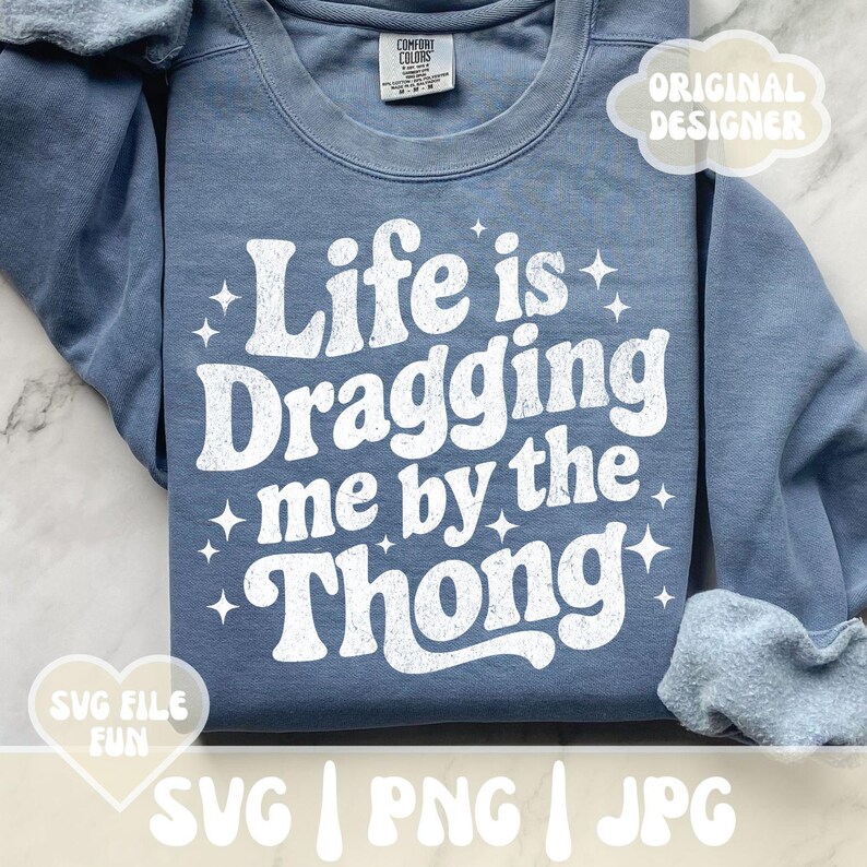 May include: A blue sweatshirt with the phrase "Life is Dragging me by the Thong" in white, retro-style lettering. The design includes star accents. The sweatshirt has a Comfort Colors tag and the words "SVG FILE FUN" and "ORIGINAL DESIGNER" are also visible.