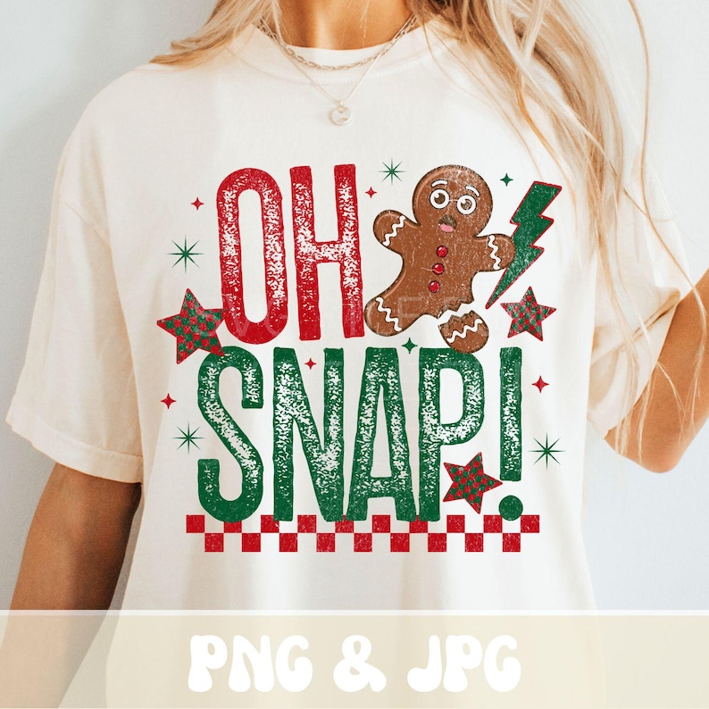 May include: Cream-colored t-shirt with a festive Christmas design. The graphic features the words "OH SNAP!" in red and green, a gingerbread man, stars, and a lightning bolt. A red and white checkered pattern is at the bottom.