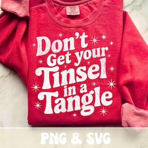 May include: Red sweatshirt with the phrase "Don't Get your Tinsel in a Tangle" in white, with star accents. The sleeves have a pinkish-red cuff. The text "PNG & SVG" is at the bottom.