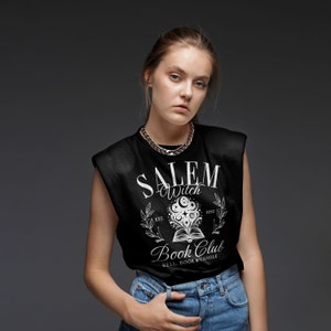 K&ouml;nnte beinhalten: Ein schwarzes &auml;rmelloses Shirt mit einem wei&szlig;en Grafikdruck, auf dem "Salem Witch Book Club" steht, mit einer hexenhaften Illustration eines Buches mit Sternen und Monden. Der Text unter der Illustration lautet "Est. 1692" und "Bell, Book & Candle".