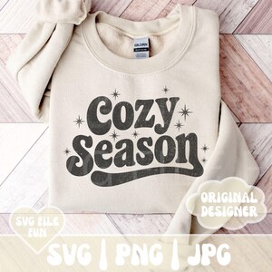 Puede incluir: Sudadera beige con la frase "Cozy Season" en una fuente retro. El texto es de color gris oscuro con detalles de estrellas. La sudadera est&aacute; doblada sobre una superficie de madera clara.